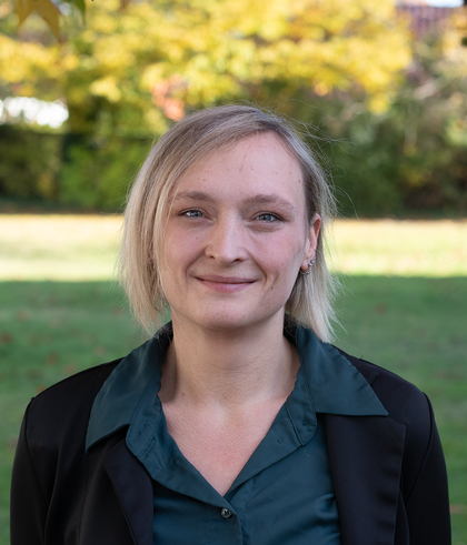 Porträt einer lächelnden Frau im Blazer vor herbstlichem Park mit unscharfem Hintergrund.