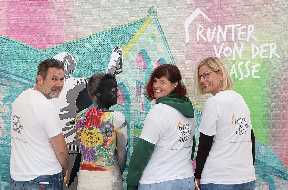Vier Personen in weißen T‑Shirts, eine zeigt eine bunte Bodypaint-Rückseite; im Hintergrund Wandgrafik mit Schriftzug „Runter von der Straße“.