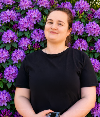 Lächelnde Frau* in schwarzem T‑Shirt vor dichtem Rhododendronbusch mit leuchtend violetten Blüten.