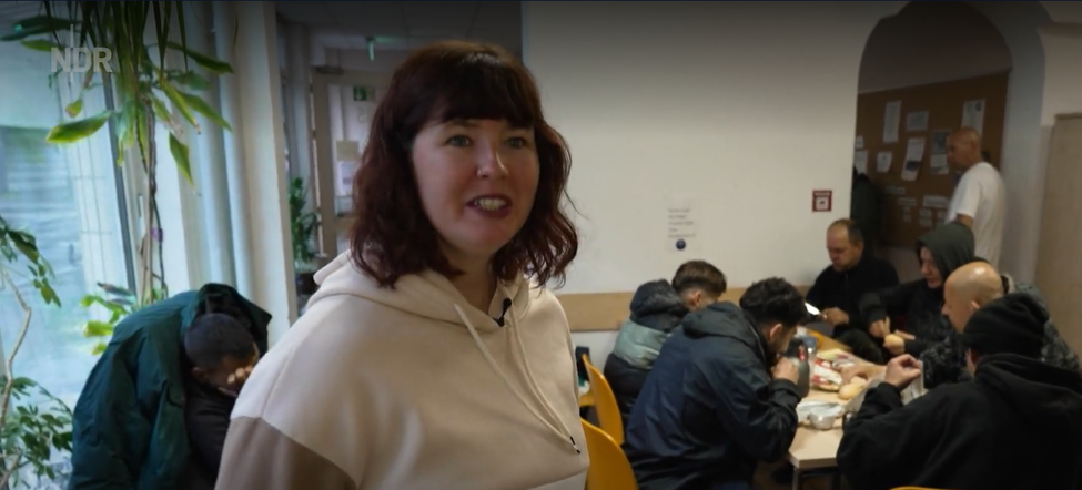Person in Kapuzenpullover im Vordergrund; im Hintergrund mehrere Menschen an Tischen, die essen und sich unterhalten.