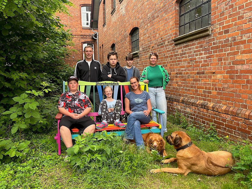 Sieben Personen sitzen und stehen an einer bunten Bank in einem Innenhof zwischen Backsteingebäuden; zwei Hunde liegen im Gras.